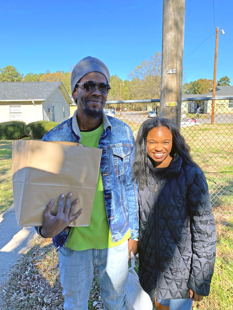 A man stands outside with a young resident and he is holding a frozen turkey in one hand and a brown paper bag in the other hand. 