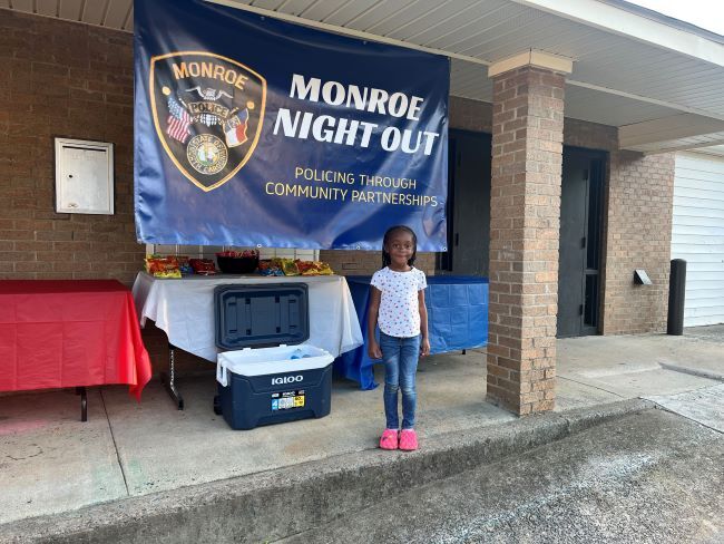 Girl standing in front of Monroe Night Out, Policing through Community Partnerships banner.